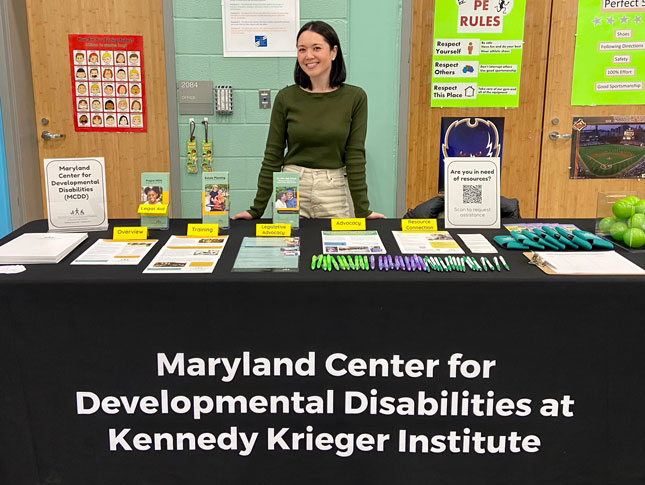 Kristine Nellenbach stands behind a table and smiles. The table is covered by a black cloth that says Maryland Center for Developmental Disabilities at Kennedy Krieger Institute.