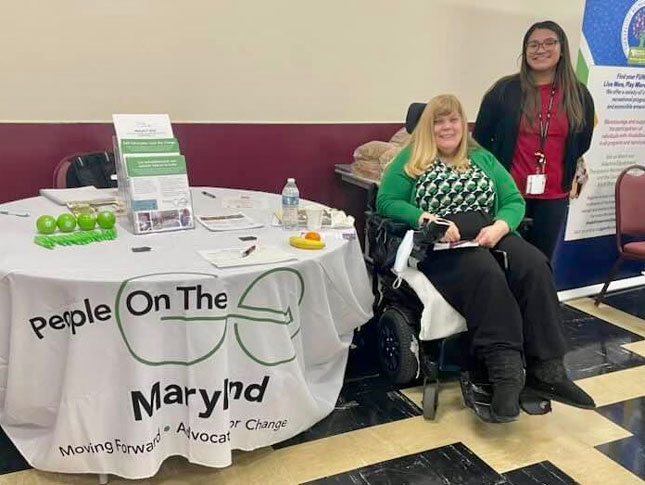 Tracy Wright and Cindy Ibarra smile, as Wright sits and her wheelchair and Ibarra stands to her left side. They are next to a table with a white People on The Go Maryland table cloth.