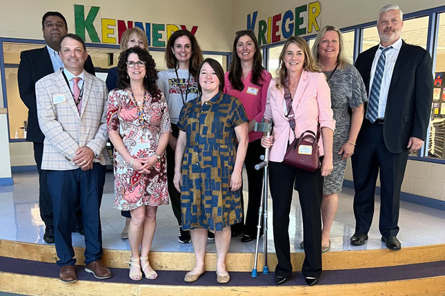 A group of 10 people in professional attire stand inside the library of Kennedy Krieger School Programs: Fairmount Campus.