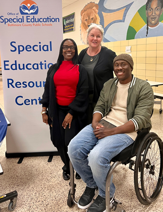 Chris Mason-Hale and two women smile in a photo at the Baltimore County Public Schools Special Education Resource Fair. He is sitting in a mobility device as the two women stand to his right.