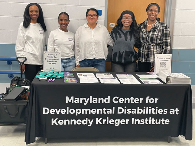 Members of the MCDD team stand behind a MCDD at Kennedy Krieger table at a resource fair.