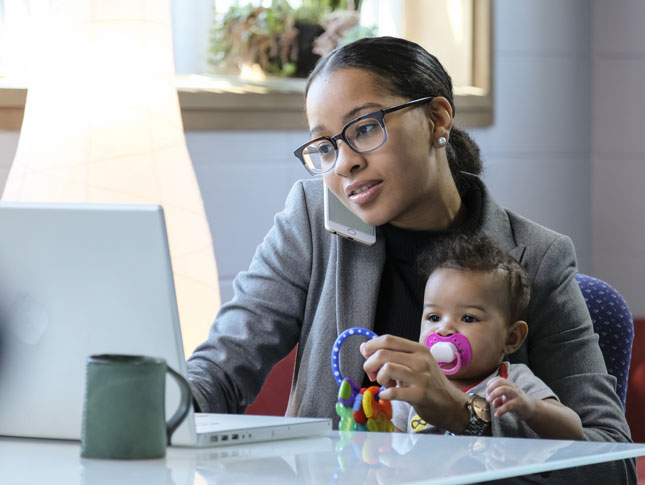 A mother talks on the phone and uses her laptop while her small child sits on her lap. 