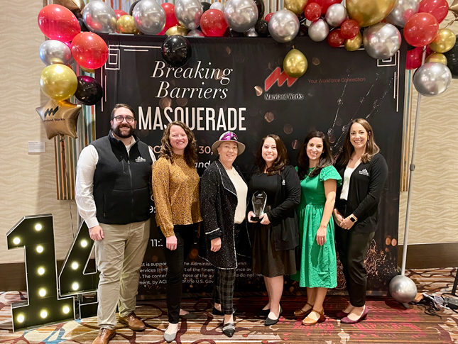 Six members of the Neurodiversity at Work Team receive the Provider of the Year award at the luncheon. The standing side by side in front of a black banner, which sits below a arch of red, gold, silver and black balloons.