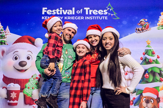 A family wearing Santa hats smiles on front of a Festival of Trees banner