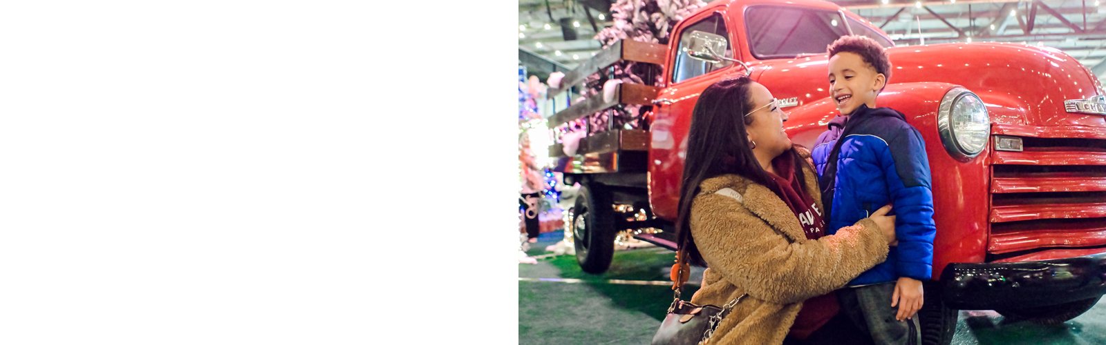 A mother and son smile while standing in front of an antique red truck on display at Festival of Trees.