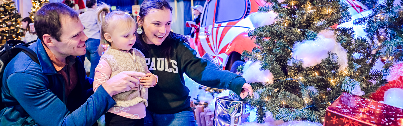 A father, mother and their young daughter smile while looking at lighted trees.