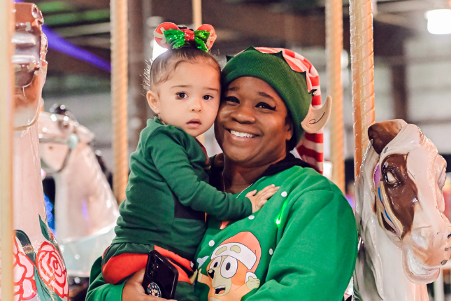 A mother holds her young daughter and smiles while standing in front of a carousel at Festival of Trees.