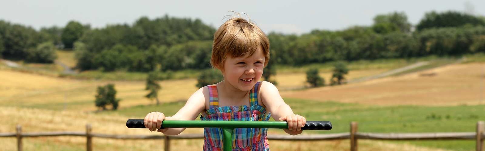 A smiling little girl with short blonde hair rides a tricycle in a rural setting.