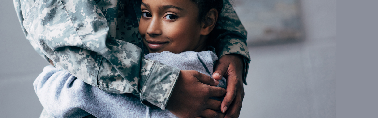 A young girl looks at the camera as she hugs an adult wearing a military uniform