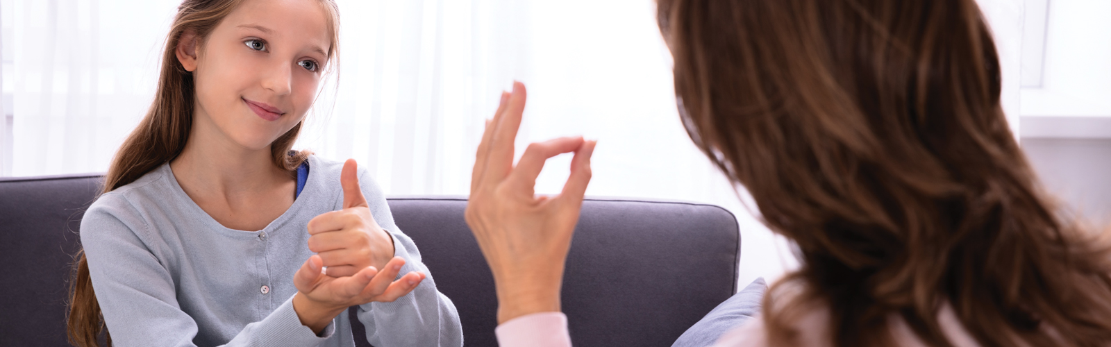 A young girl sits on a sofa communicating in sign language from an adult woman sitting across from her. The adult is seen from behind.