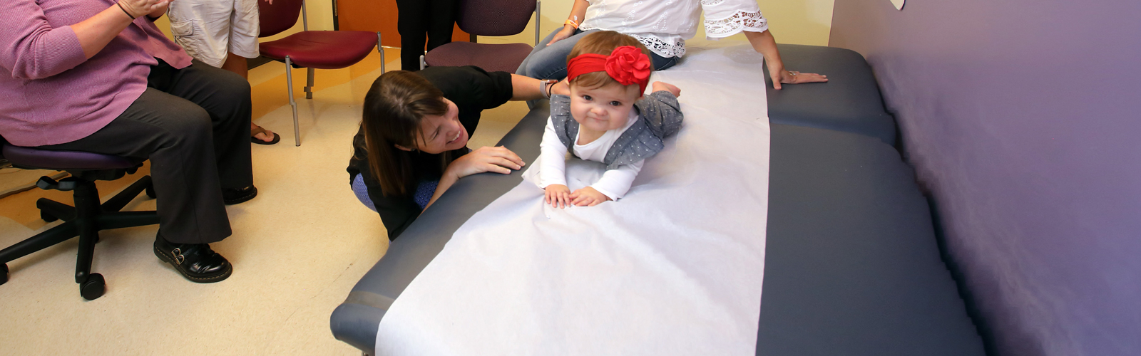 A baby lays on her stomach on an examination table, while her doctor supports her back. The doctor is smiling as she supports the baby.