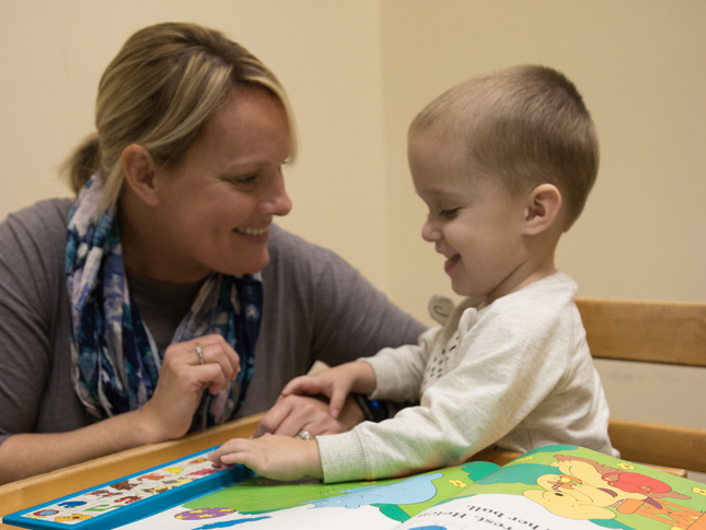 A speech-language therapist works with a young boy as he plays with toys on a table. She is kneeling down to his level and both are smiling.
