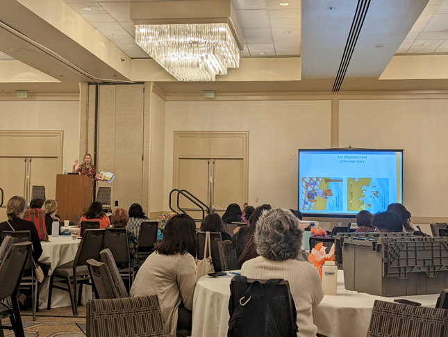 Dr. Rebecca Landa gives a presentation during the 21st Annual Autism Conference. She is standing at a podium as people sit at tables in front of her watching.  A  projection screen on the right  shows the presentation. 