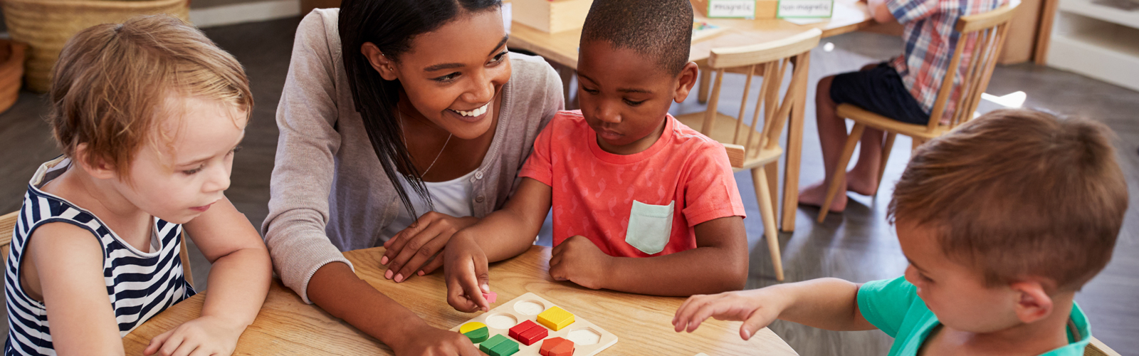 A teacher smiles as she helps three pre-school students with an activity at their desk.