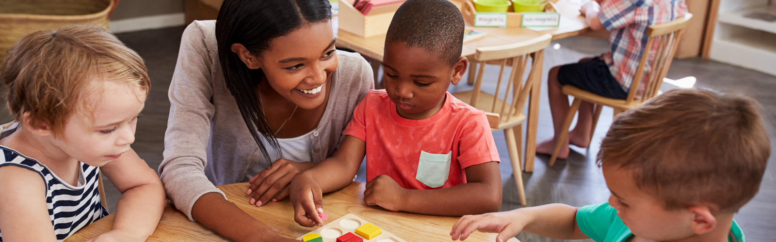 A teacher smiles as she helps three pre-school students with an activity at their desk.