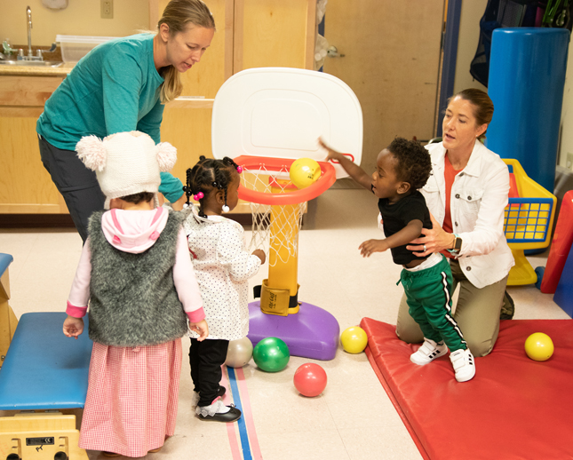 Therapists assist children in the Happy Feet group with getting balls through a basketball hoop.