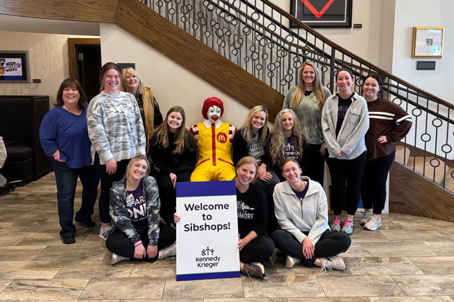 A group of people pose for a photo around a statue of Ronald McDonald sitting on a bench at Ronald McDonald House. A person sitting in front of the statue is holding up a Kennedy Krieger branded sign that says Welcome to Sibshops!