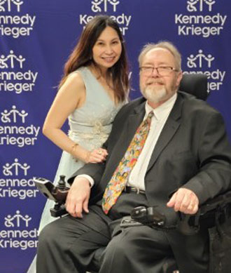 A woman stands next to a man in a wheelchair at ICSCI's 20th anniversary Gala. Both are smiling as stand in front of a Kennedy Krieger-branded step and repeat banner.