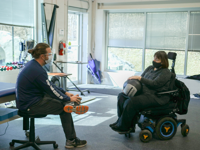 A woman sitting in a wheelchair rests a medicine ball in her lap during a personal training session with her therapist.