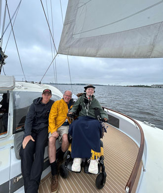 Two men and a woman smile while aboard the Impossible Dream in the Inner Harbor. The two men are seated on a bench while the woman is using her wheelchair.