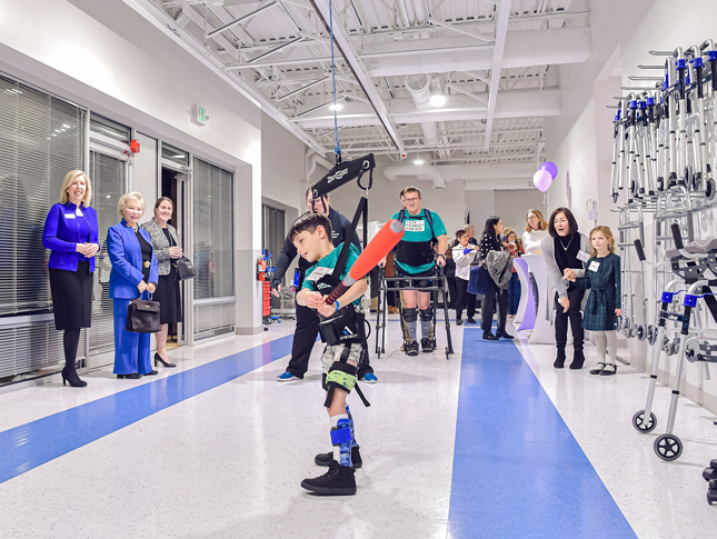 A room of grand opening attendees watch as a boy swings a foam baseball bat while using the a ZeroG 3D support system.