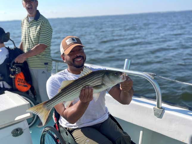 A man in a wheelchair holds a fish during an Adaptive Sports fishing trip. He is smiling on a boat, and the water can be seen in the background.