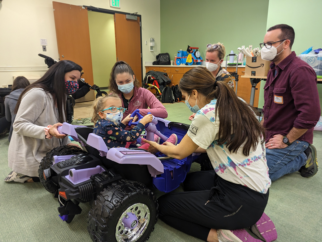 A young girl sits in a modify ride-on toy while being assisted by three ICSCI staff members. Two other adults look on from the front of the toy.