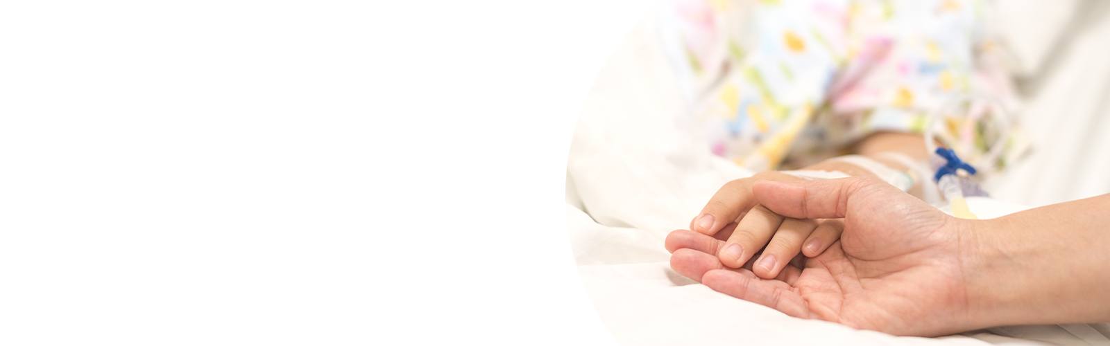 Close-up of a child lying in a hospital bed holding an adult’s hand, with an IV line visible.
