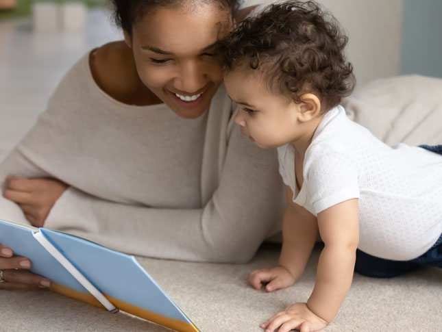 A mother reads to her young toddler as they lay on their stomachs. 
