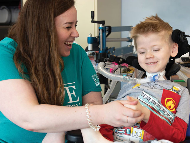 A young boy in a wheelchair smiles while holding his therapist's hand. The therapist is to his right, and she's looking at him and smiling.