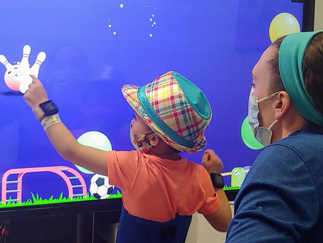 A young boy plays a touch screen game as part of his therapy, with a therapist kneeling behind him.