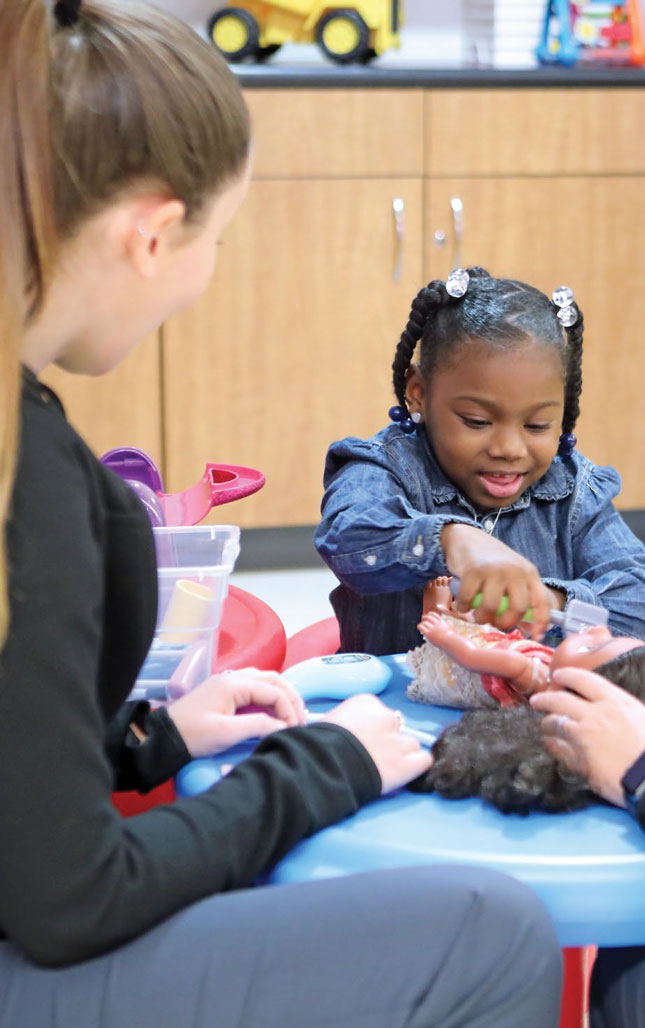 A young girl smiles while performing an activity. A therapist sits to her right with her back to the camera.