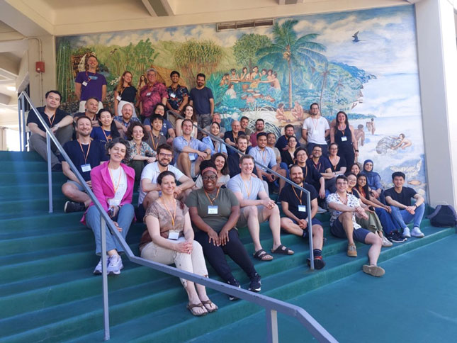 Attendees of the 3rd installation of the MRS Hackathon in a group photo. The photo is staged on a staircase. Alongside the staircase, there is a colorful mural depicting a beach scene.