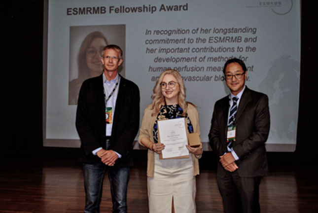 Dr. Linda Knutsson holds a certificate recognizing her fellowship of the European Society for Magnetic Resonance in Medicine and Biology (ESMRMB). She is standing between two men, with a slide summarizing her accomplishments on a screen behind them.