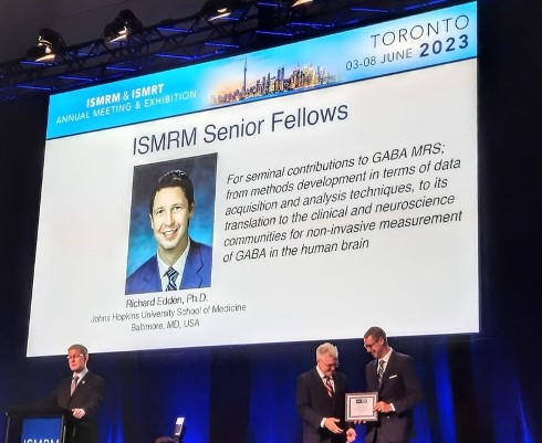 Dr. Richard Edden is honored as senior Fellow of the International Society for Magnetic Resonance in Medicine (ISMRM) at its annual meeting in Toronto, Canada. He is standing on stage receiving an award, while a slide shows his headshot and a brief bio on projection screen behind him.