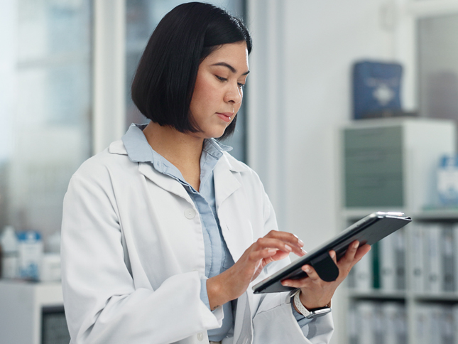 A woman wearing a lab coat types research data into a tablet while standing in a research lab.