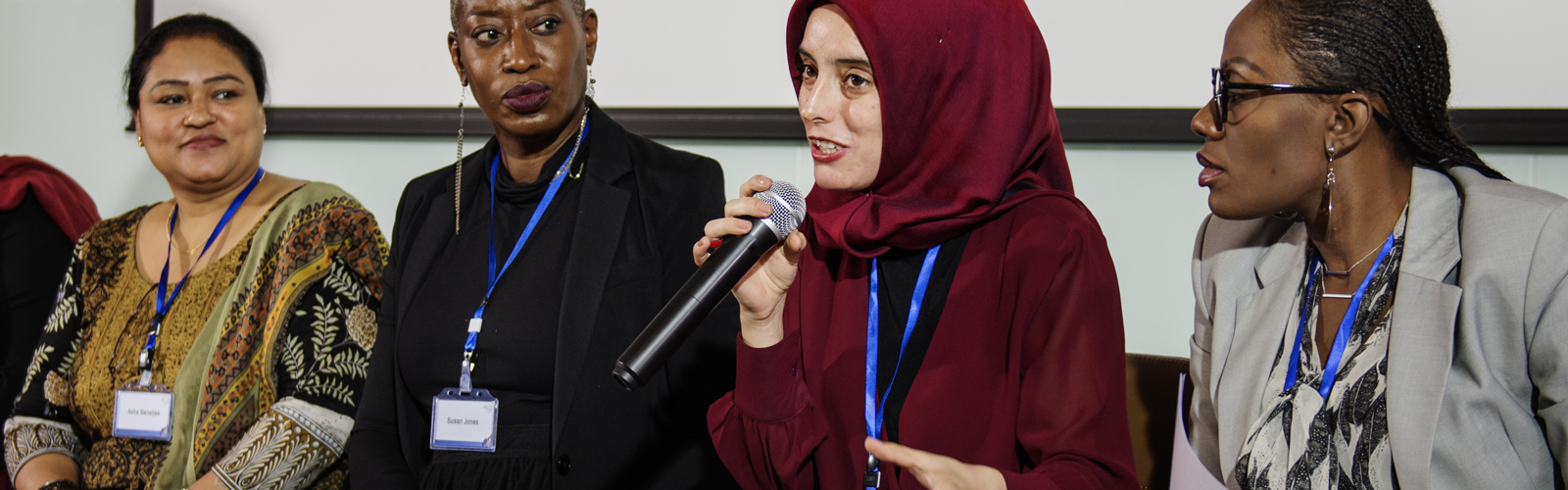 Four women participate in a panel discussion at a professional event. One of them is holding a microphone and speaking as the others listen intently. 