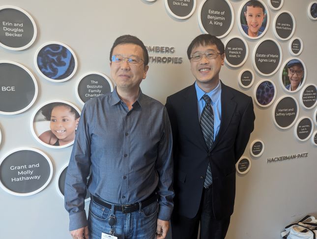 Two men stand next to each other and smile. They are standing in front of a wall that recognizes the names of donors to Kennedy Krieger Institute in circular signs.