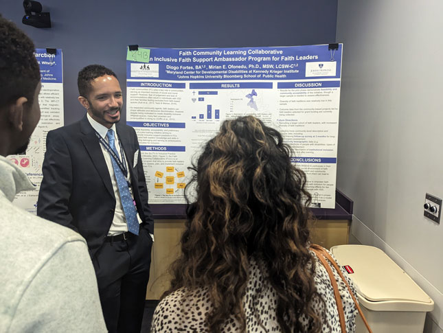 A man stands in front of a research poster and speaks to two people. The other two individuals have their backs to the camera.