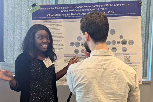 A woman smiles while talking to a man as they stand in front of a research poster. She is facing the camera, while his back is to the camera.