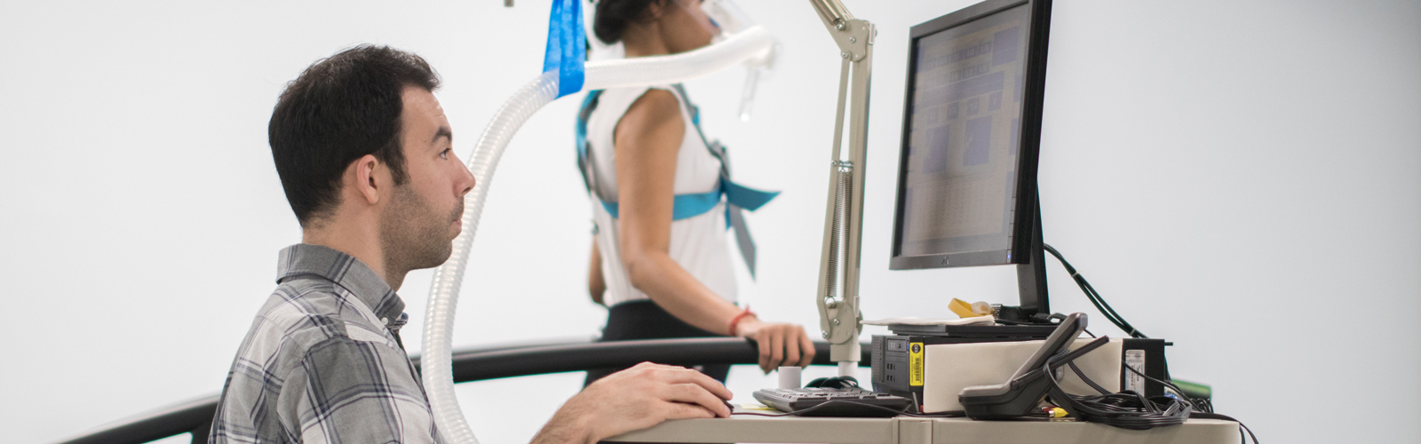 A study participant walks on a research treadmill while a researcher stands to their right side monitoring data on a computer screen.