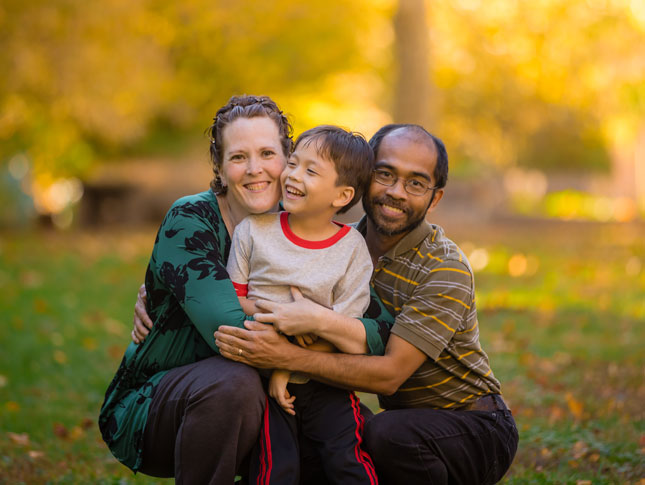 A mother and father smile with their son in a photo. The mother and father are kneeling down while their son leans against them. 