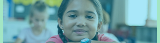 A young girl looks into the camera while sitting at her desk in a classroom.