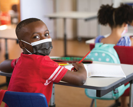 A young boy at his school desk looks over his shoulder at the camera while holding a pencil in his right hand and wearing a face mask