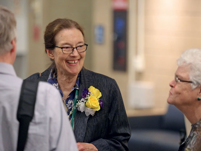 A woman smiles and looks off to the side. She is wearing a corsage of yellow roses and purple flowers, with a white bow. In the foreground is a man’s blurry back. They are standing in a room or hallway.