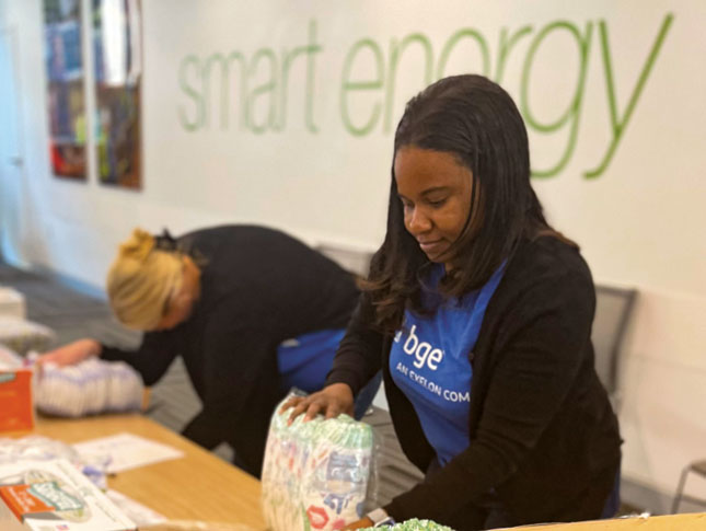 Two women work at a table assembling diapers into care packages. Each woman is wearing a blue T-shirt. One of the T-shirts can be seen to read “BGE, an Exelon Company.”