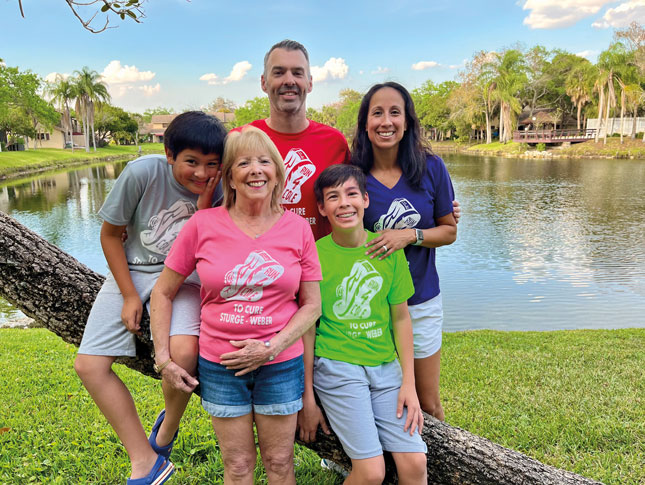 Portrait photo of a family outside, with a pond and palm trees in the background. The family members are Mom, Dad, Grandma and two boys. Each person wears a T-shirt with “Run 4 Cole to cure Sturge-Weber” on it. Each T-shirt is a different color.