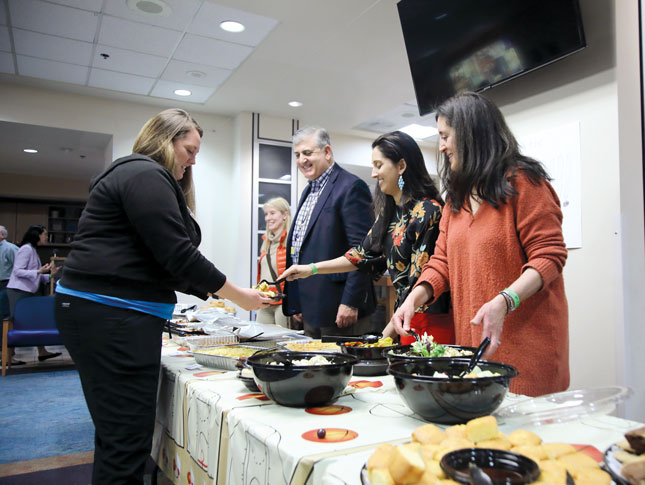 Four people stand on one side of a table, serving food. On the other side, a woman receives a plate of food.