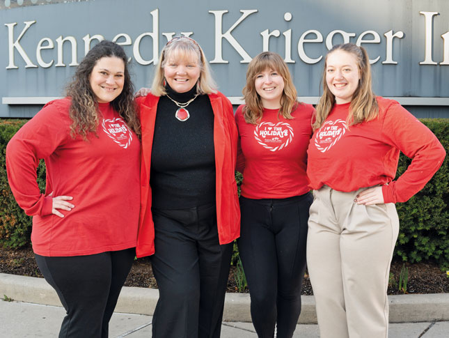 Four women stand outside in front of a sign that says “Kennedy Krieger.” Three of them are wearing red T-shirts that say “I love the holidays,” with the Kennedy Krieger Festival of Trees logo underneath.