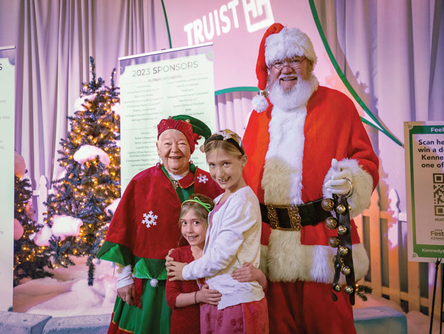 Santa and Mrs. Claus pose with two girls. All four people are white, and they are standing in an indoor space that looks like it could be a convention hall. Behind them are artificial Christmas trees covered in white lights and artificial snow. Also behind them is a pull-down banner that reads, “2023 SPONSORS,” followed by a list that is not readable in the photo. Behind the banner is a sign that reads, “TRUIST,” with a logo after it.